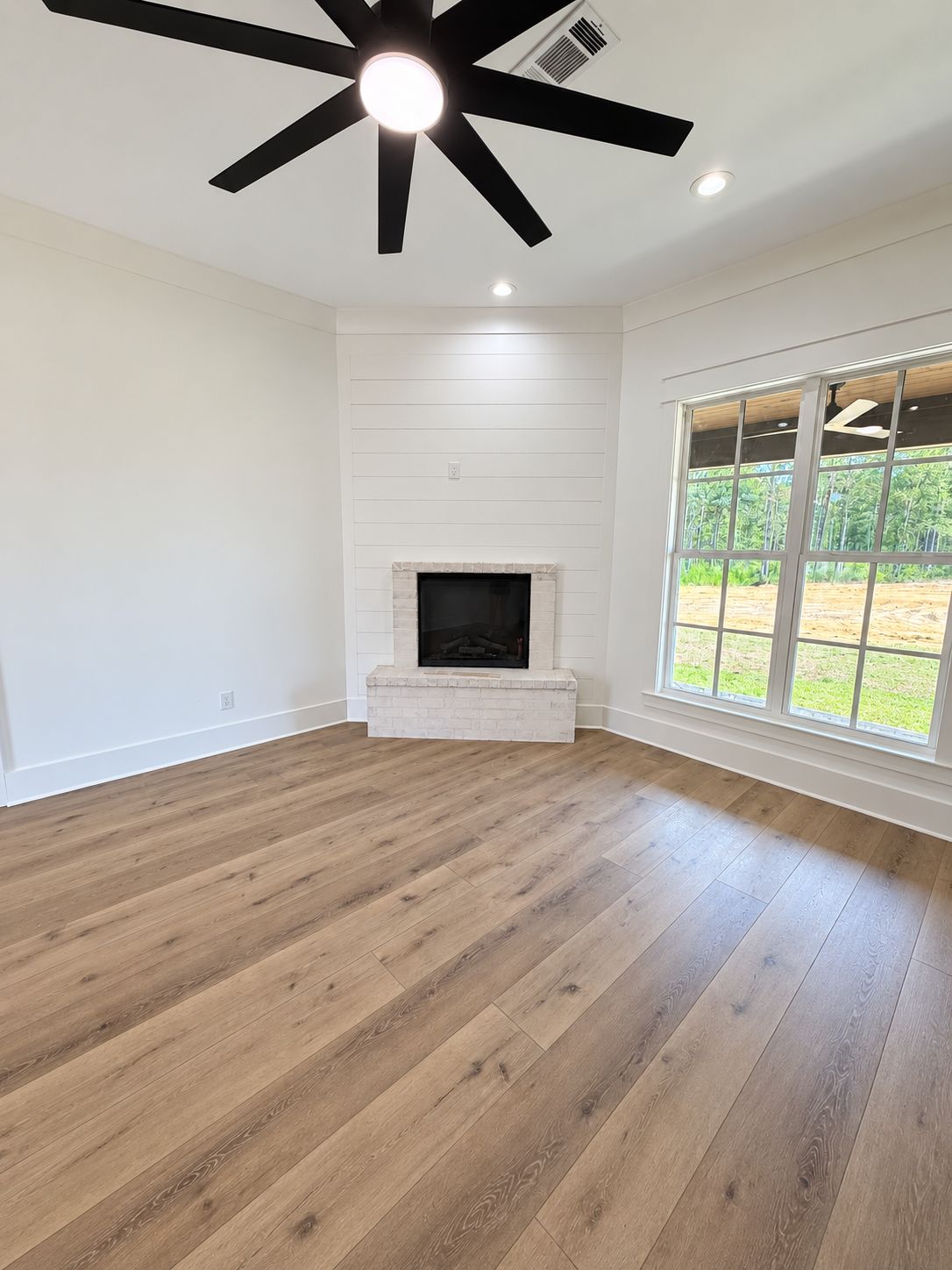 Living room with a white brick fireplace, large windows, and a black ceiling fan.