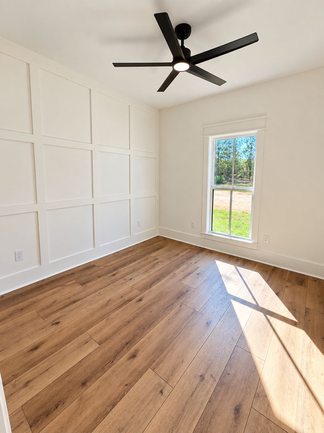 Room with one tall window, a black ceiling fan, and white paneled walls.