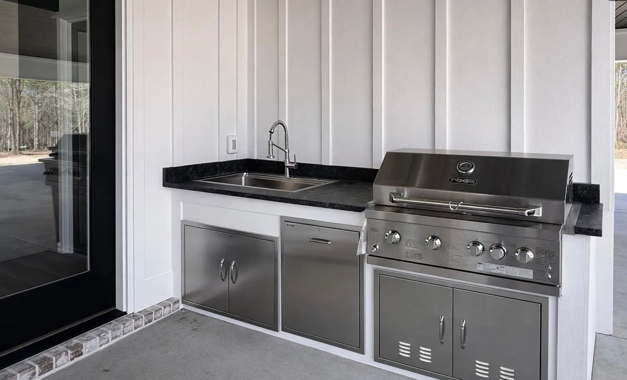 Covered outdoor kitchen with stainless steel cabinets, a sink, grill, and black countertops.